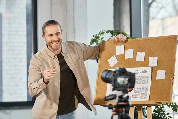 Возбужденный бизнесмен показывает биткойн на цифровой камере рядом с пробковыми досками с графиками и бумажными заметками — стоковое фото
