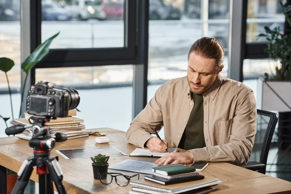 Homem de negócios sério em traje casual escrevendo em notebook na mesa de trabalho na frente da câmera digital — Fotografia de Stock