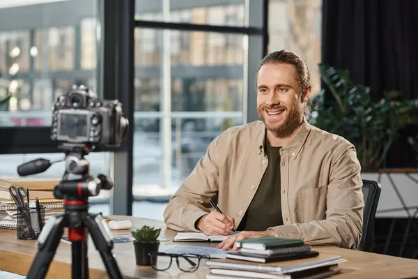 Homme d'affaires heureux souriant à l'appareil photo numérique et l'écriture dans un cahier sur le lieu de travail dans le bureau moderne — Photo de stock
