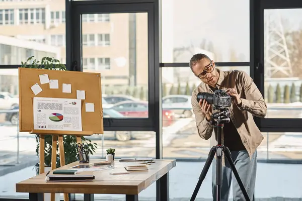 Hombre de negocios en gafas ajustando cámara digital cerca de escritorio y corcho con gráficos en la oficina - foto de stock