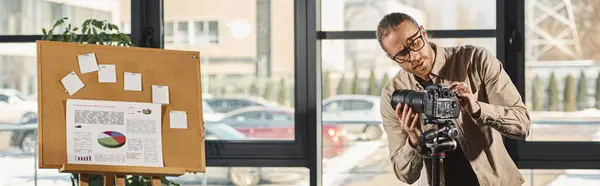 Manager en tenue décontractée et lunettes ajustant appareil photo numérique à côté de carton-liège avec graphiques, bannière — Photo de stock