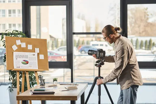 Предприниматель в повседневной одежде регулирует цифровую камеру возле стола и пробковую доску с графиками в офисе — стоковое фото