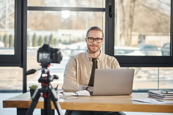 Glücklicher Manager in Brille sitzt neben Laptop und lächelt während Videoblog in die Digitalkamera — Stockfoto