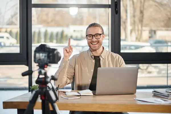 Glücklicher Geschäftsmann zeigt Ideenschild, während er neben Laptop vor Digitalkamera sitzt, Videoblog — Stockfoto