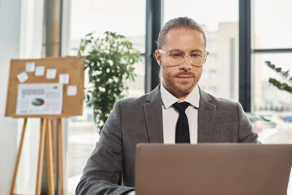 Manager fiducioso in occhiali e tuta grigia che lavora su laptop in un ambiente ufficio moderno — Foto stock