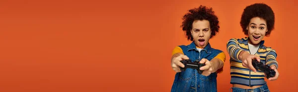 Cheerful young african american brother and sister playing video games with joysticks, banner — Stock Photo