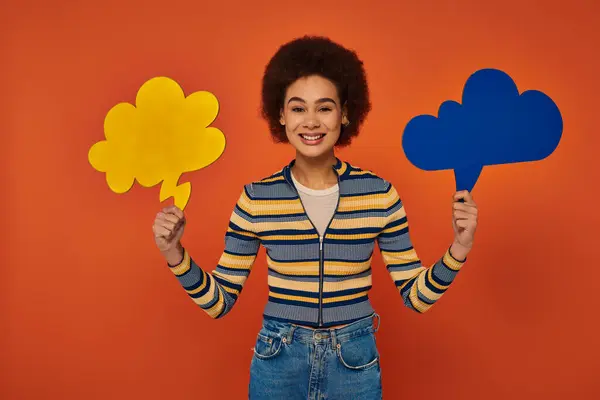 Jovem mulher afro-americana posando com bolhas de pensamento azul e amarelo no fundo laranja — Fotografia de Stock