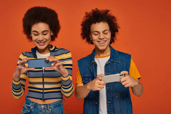 Fröhliche afrikanisch-amerikanische Schwester und Bruder schauen glücklich auf ihre Mobiltelefone, orangefarbene Hintergrund — Stockfoto