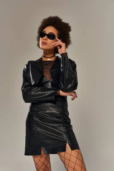 Attractive stylish african american woman in black attire with sunglasses posing on gray backdrop — Stock Photo
