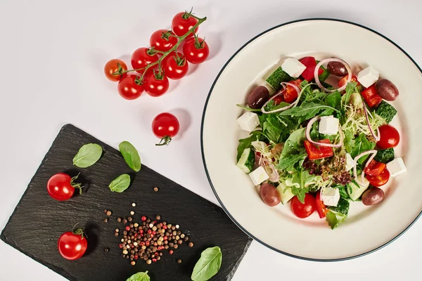 Фото тарелки с традиционным греческим салатом рядом с помидорами и листьями базилика — стоковое фото