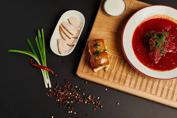 Свіжий український буряковий суп біля часникових булочок, свинячого салату та зеленої цибулі на обробній дошці на чорному — стокове фото
