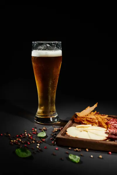 Copo de cerveja artesanal perto de papelão charcutaria com queijo gourmet, carne seca e salame em preto — Fotografia de Stock