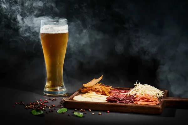 Vaso de cerveza artesanal cerca del tablero de charcutería con selección de queso, carne seca y salami en negro - foto de stock