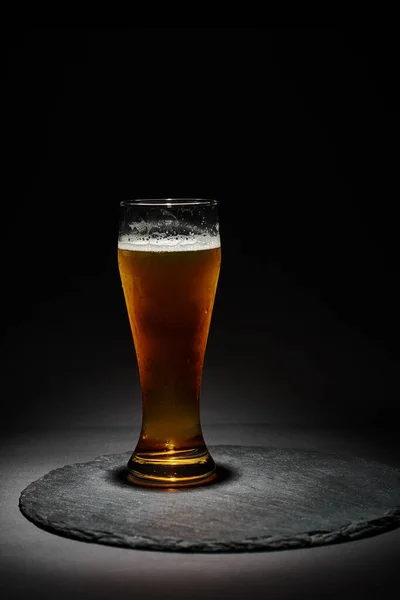 Vidro de cerveja artesanal ou lager com espuma em vidro resfriado na montanha-russa de ardósia em fundo preto — Fotografia de Stock
