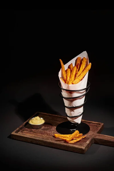 Tasty crispy French fries inside of paper cone near dipping sauce on wooden cutting board — Stock Photo