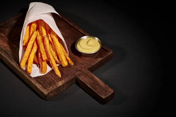 Salty and crispy French fries inside of paper cone near dipping sauce on wooden cutting board — Stock Photo