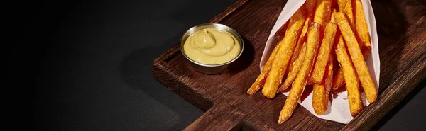 Banner of salty crispy French fries inside of paper cone near dipping sauce on wooden cutting board — Stock Photo