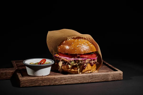 Delizioso cheeseburger con panino al sesamo, manzo e sottaceti come contorno su vassoio di legno su nero — Foto stock