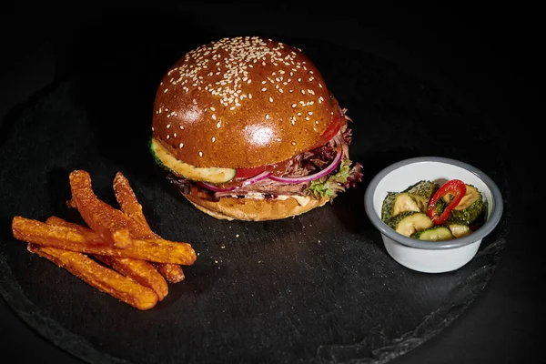 Hamburger savoureux avec du bœuf à l'intérieur de pain de sésame près des cornichons comme plat d'accompagnement sur fond noir — Photo de stock