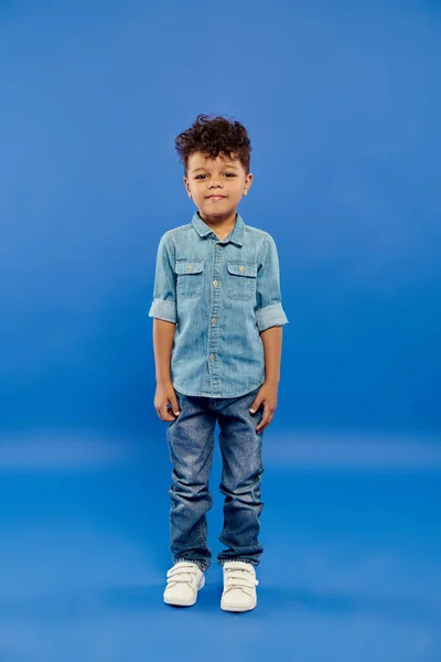 Cute and curly african american preschooler boy in stylish denim outfit posing on blue background — Stock Photo