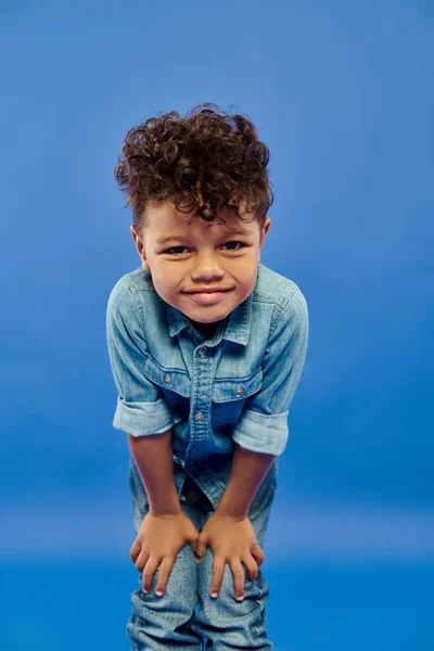 Pequeno menino pré-escolar afro-americano feliz em roupa de ganga elegante posando no fundo azul — Fotografia de Stock