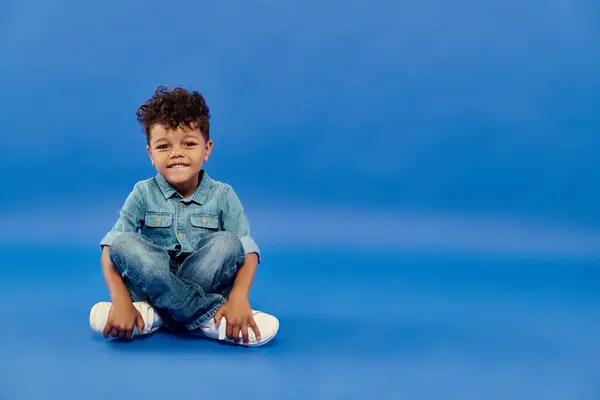 Happy curly african american preschooler boy in stylish denim outfit sitting on blue background — Stock Photo
