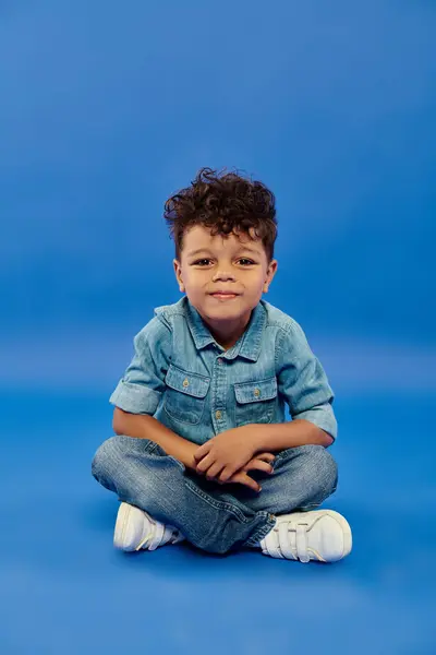 Sorrindo encaracolado menino pré-escolar afro-americano em roupa de ganga elegante sentado no fundo azul — Fotografia de Stock