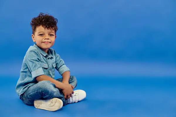 Sorrindo e bonito menino pré-escolar afro-americano em roupa de ganga elegante sentado no fundo azul — Fotografia de Stock
