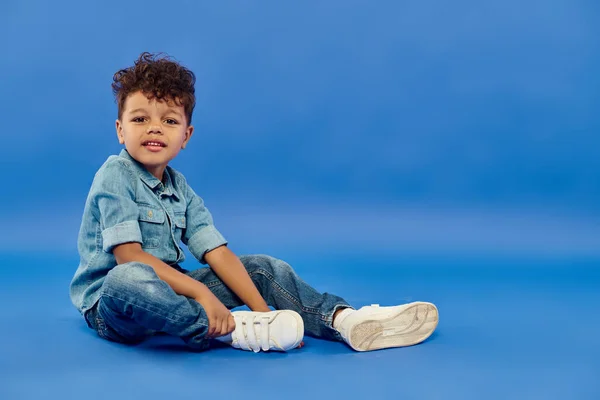 Smiling african american preschooler boy in stylish denim attire sitting on blue background — Stock Photo