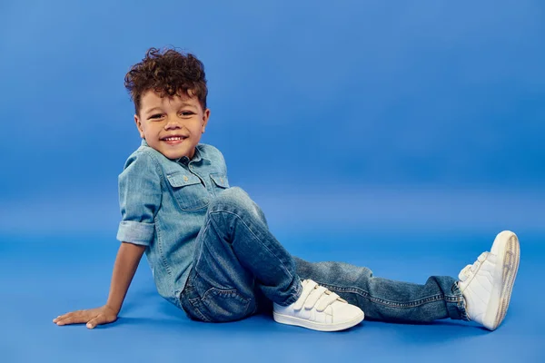 Positive african american preschooler boy in stylish denim attire sitting on blue background — Stock Photo