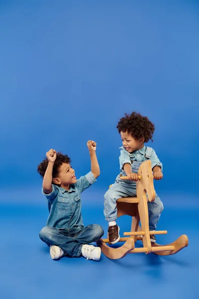 Feliz menino criança afro-americano em roupas de ganga elegante sentado no cavalo de balanço perto do irmão — Fotografia de Stock