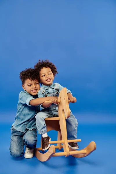 Alegre afro-americano criança menino em roupas de ganga elegante sentado no cavalo de balanço perto do irmão — Fotografia de Stock