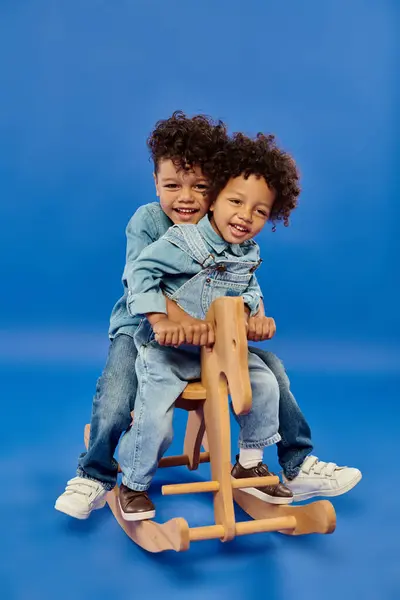 Heureux afro-américains garçons en jean élégant vêtements assis sur cheval à bascule sur fond bleu — Photo de stock