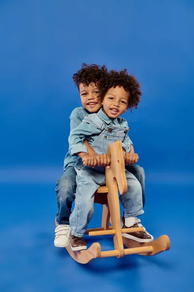 Feliz afroamericano hermanos en denim ropa sentado juntos en balanceo caballo en azul telón de fondo - foto de stock