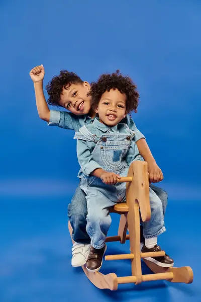 Crianças americanas africanas felizes em roupas jeans sentadas juntas no cavalo de balanço em pano de fundo azul — Fotografia de Stock