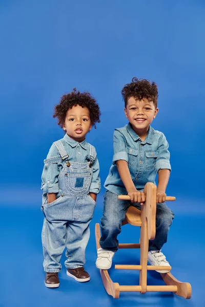 Mignon jeune garçon afro-américain en denim élégant vêtements debout près du frère sur cheval à bascule — Photo de stock