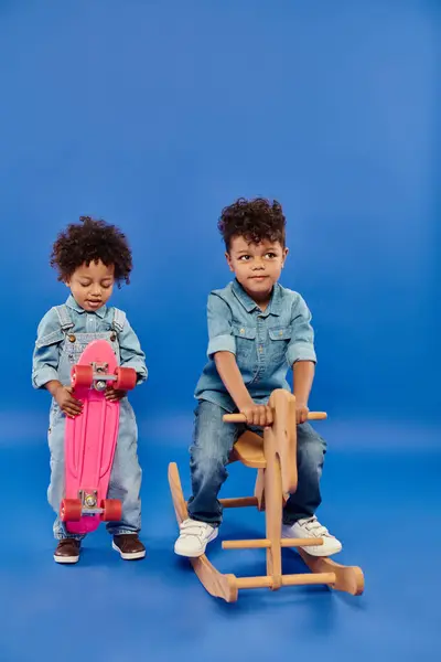 African american boy in stylish denim clothes holding penny board near brother on rocking horse — Stock Photo