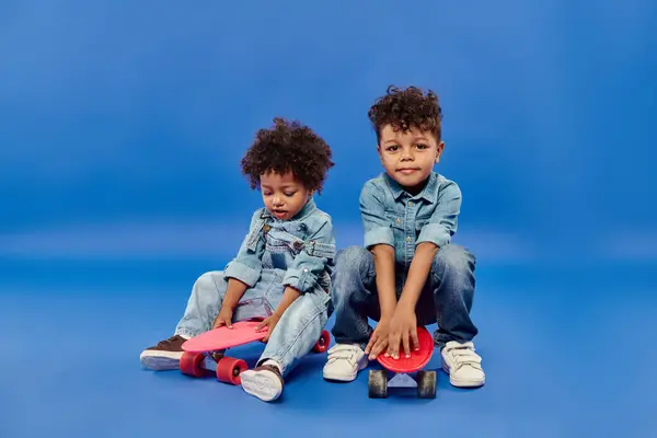 Симпатичные африканские американские братья и сестры в стильной джинсовой одежде, сидящие на пенни досках на синем фоне — стоковое фото