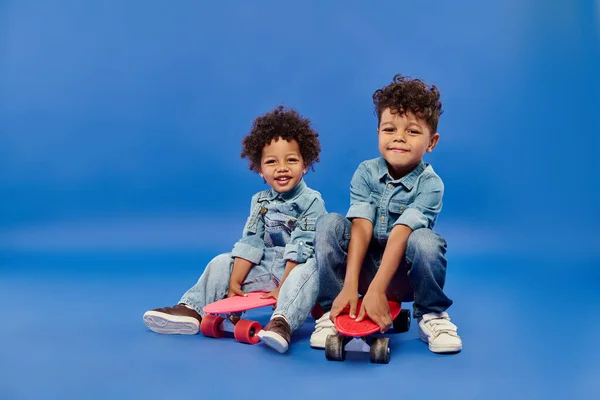 Heureux frères et sœurs afro-américains dans des vêtements en denim élégant assis sur des planches de penny sur fond bleu — Photo de stock