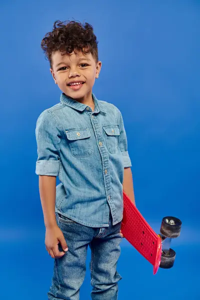 Heureux garçon préscolaire afro-américain en denim élégant vêtements debout sur le panneau penny sur bleu — Photo de stock