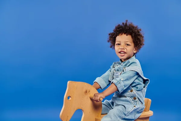 Happy african american toddler boy in denim clothes sitting on rocking chair on blue backdrop — Stock Photo