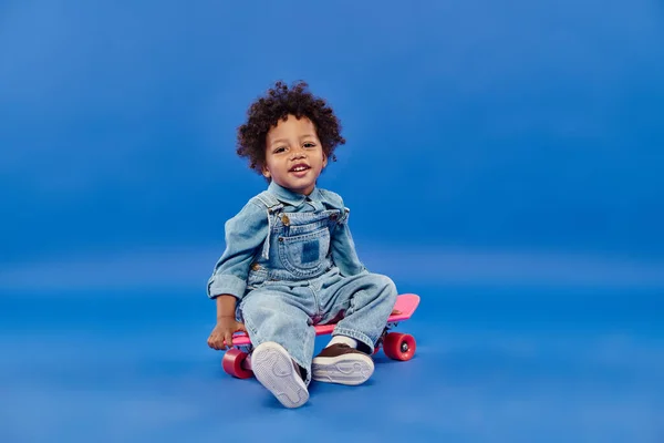 Fröhliche afrikanisch-amerikanische Kleinkind Junge in Jeanskleidung sitzt auf Penny Board auf blauem Hintergrund — Stockfoto