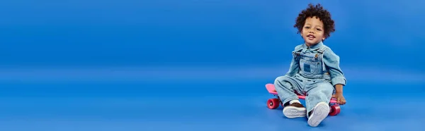 Happy african american toddler boy in denim clothes sitting on penny board on blue backdrop, banner — Stock Photo