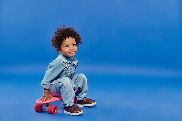 Adorable afroamericano niño en ropa de mezclilla sentado en penny board sobre fondo azul - foto de stock