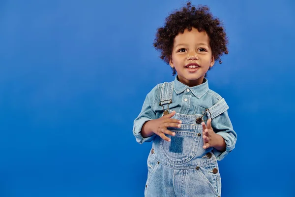 Adorável e encaracolado afro-americano criança menino em roupas de ganga elegante em pé no fundo azul — Fotografia de Stock