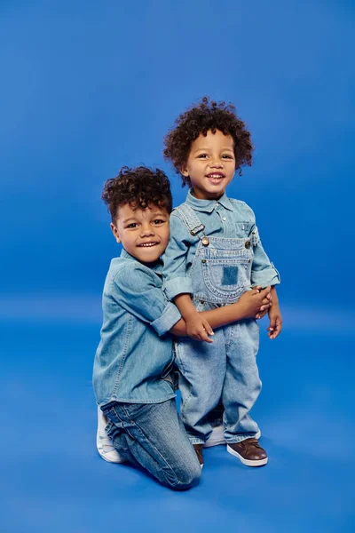 Happy african american preschooler boy in denim clothes embracing toddler brother on blue background — Stock Photo
