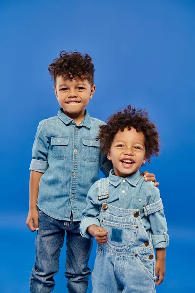 Joyful african american preschooler boy in denim clothes standing with toddler brother on blue — Stock Photo