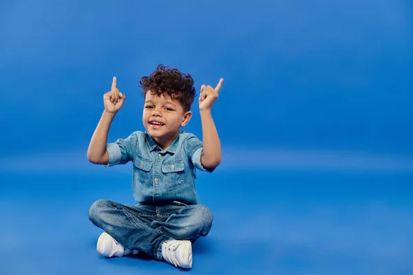 Joyful and curly african american preschooler boy in denim clothes sitting pointing up on blue — Stock Photo