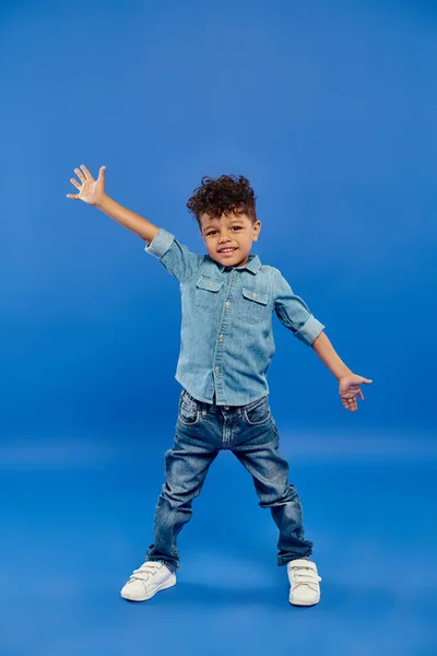 Joyful and curly african american preschooler boy in denim clothes standing and gesturing on blue — Stock Photo