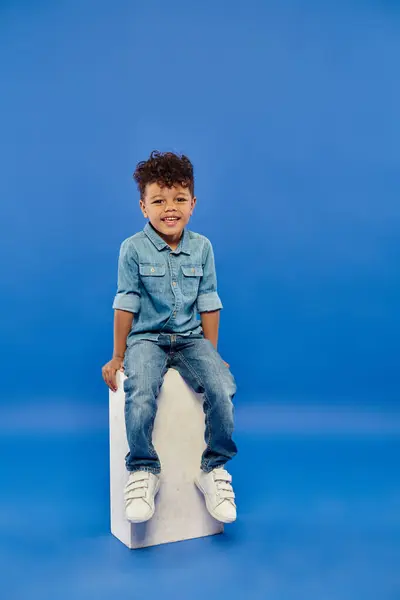 Niño preescolar afroamericano feliz y rizado en ropa de mezclilla sentado en cubo blanco en azul - foto de stock